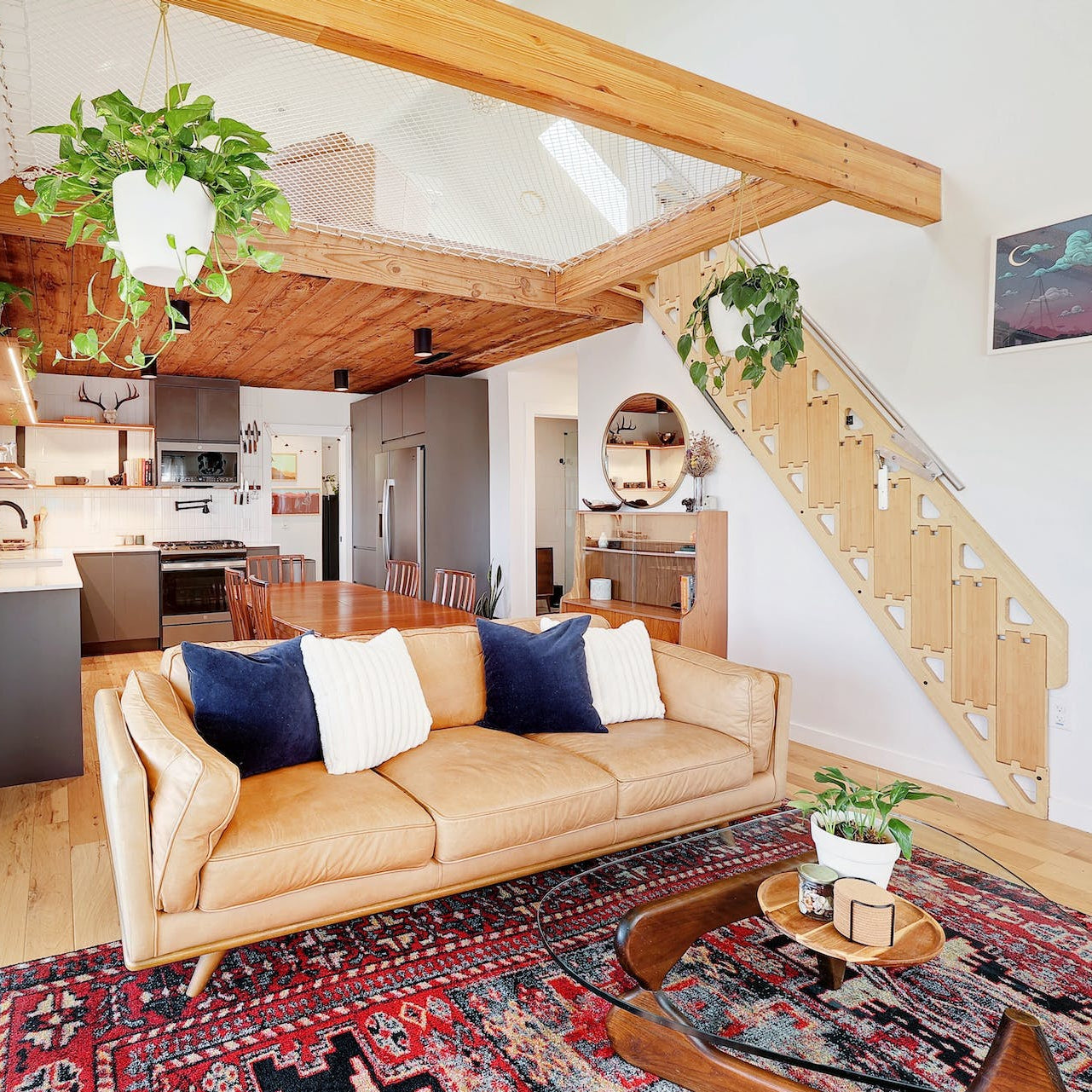 A modern open-plan living space featuring a light tan leather sofa with navy and white pillows, a red patterned rug, a wooden staircase, and a kitchen in the background.